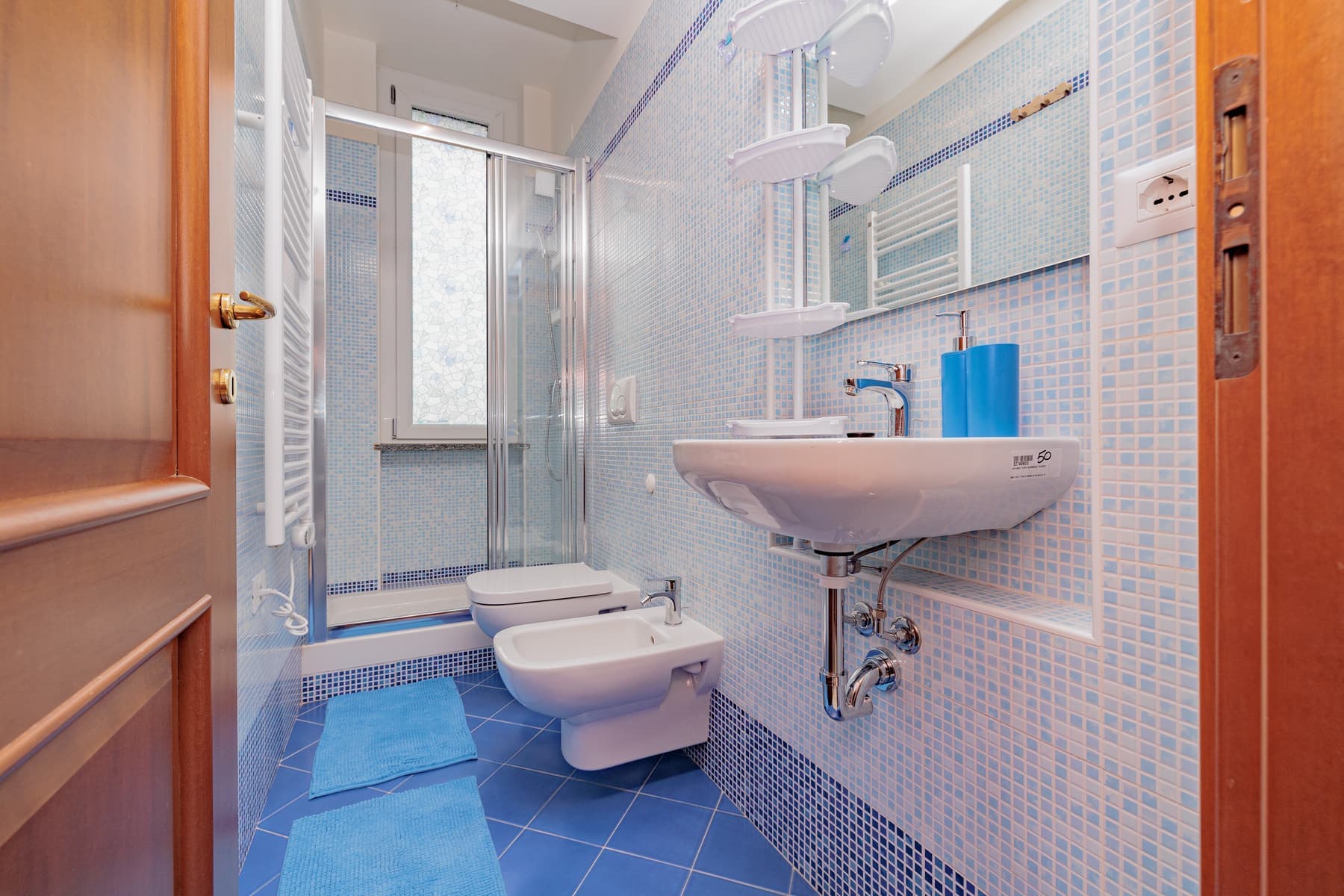 Mosaico azzurro e sanitari sospesi: Bagno rivestito a mosaico su tutte le pareti, con box doccia e sanitari sospesi.