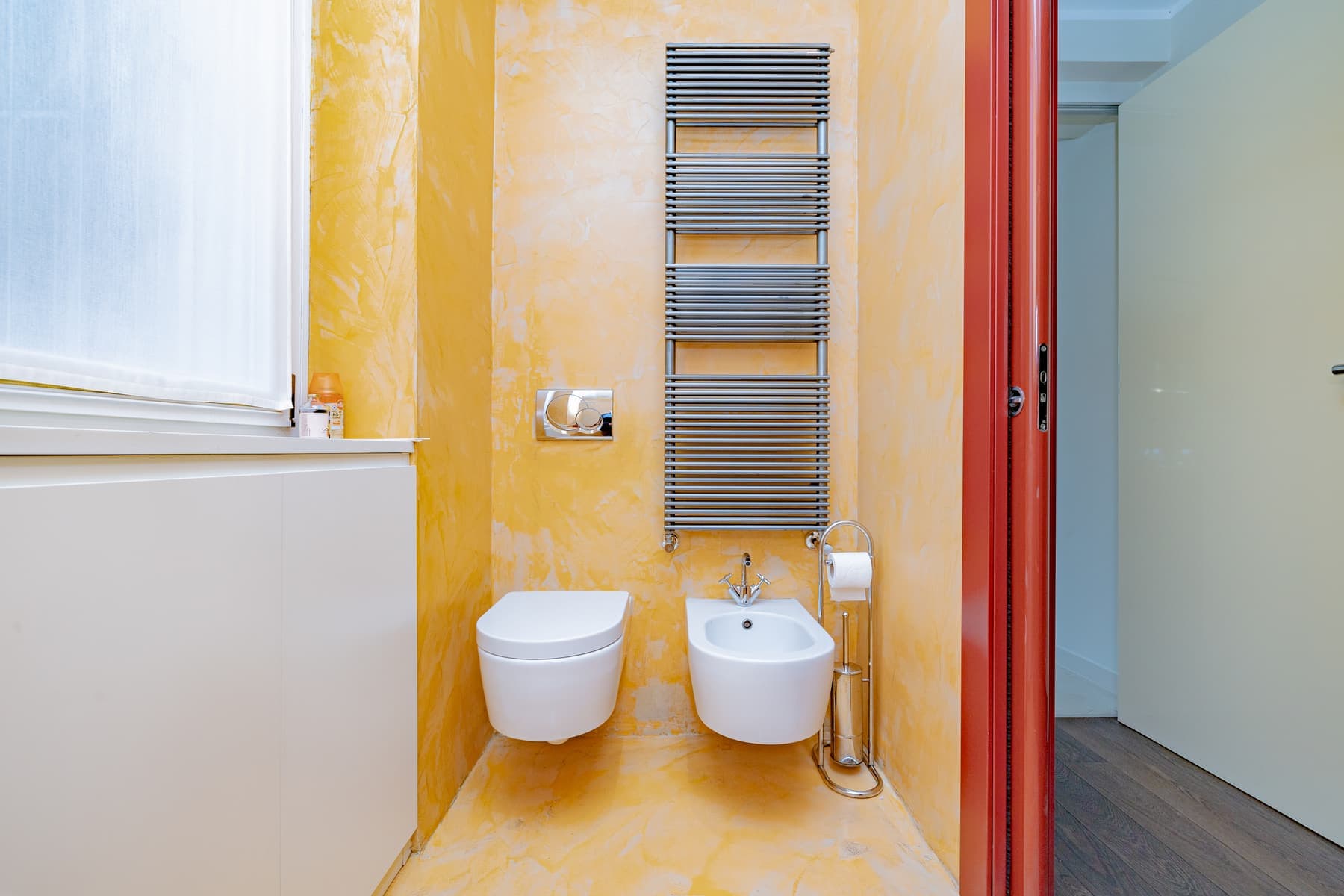 Spatolato giallo e porta rossa: Bagno di servizio con pareti a spatolato, sanitari sospesi e scaldasalviette cromato.