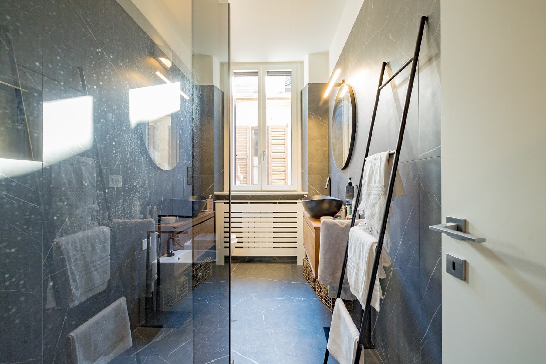 Grandi lastre grigie e lavabo nero: Pareti in grandi lastre effetto pietra, lavabo d'appoggio nero su piano in legno e specchio tondo.